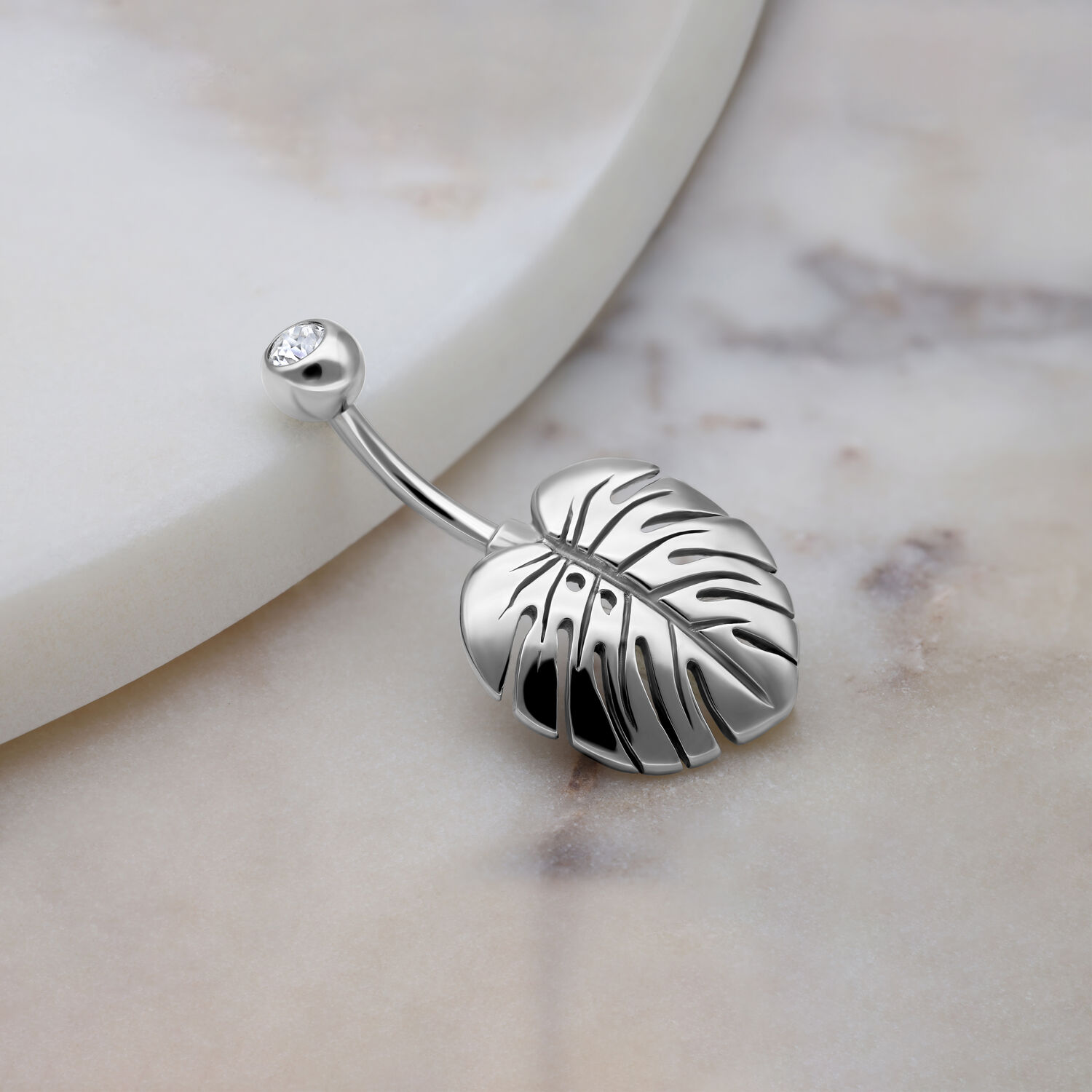Silbernes Bauchnabelpiercing mit Monstera Blatt und Kristall auf Marmorhintergrund