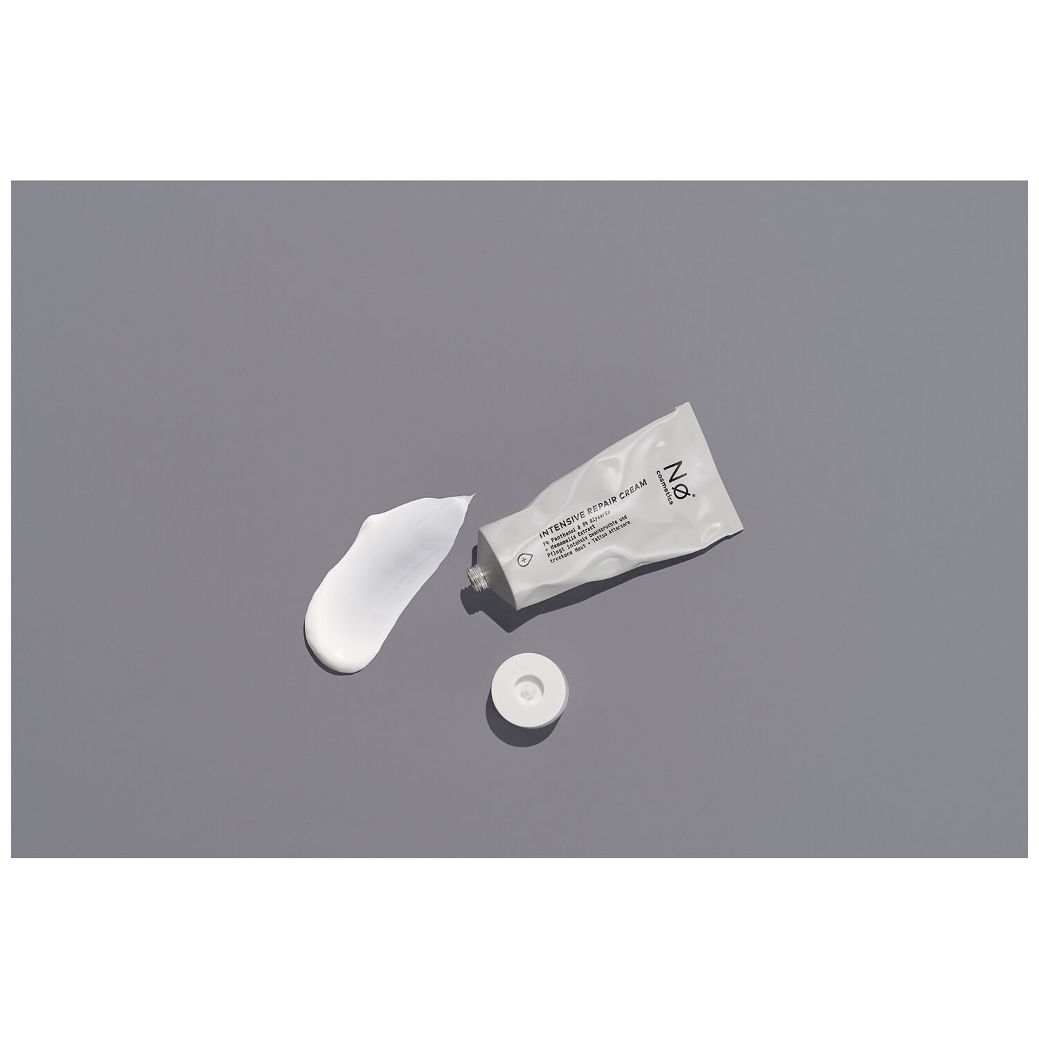 Graue Tube der Nø Cosmetics Intensiv Repair Cream mit abgenommenem Deckel und etwas Creme daneben auf grauem Hintergrund