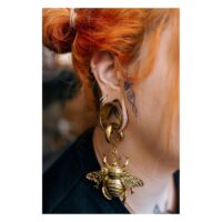 Frau mit rotorangenen Haaren trägt goldenen Ohrring mit großem Bienenanhänger vor verschwommenem Hintergrund
