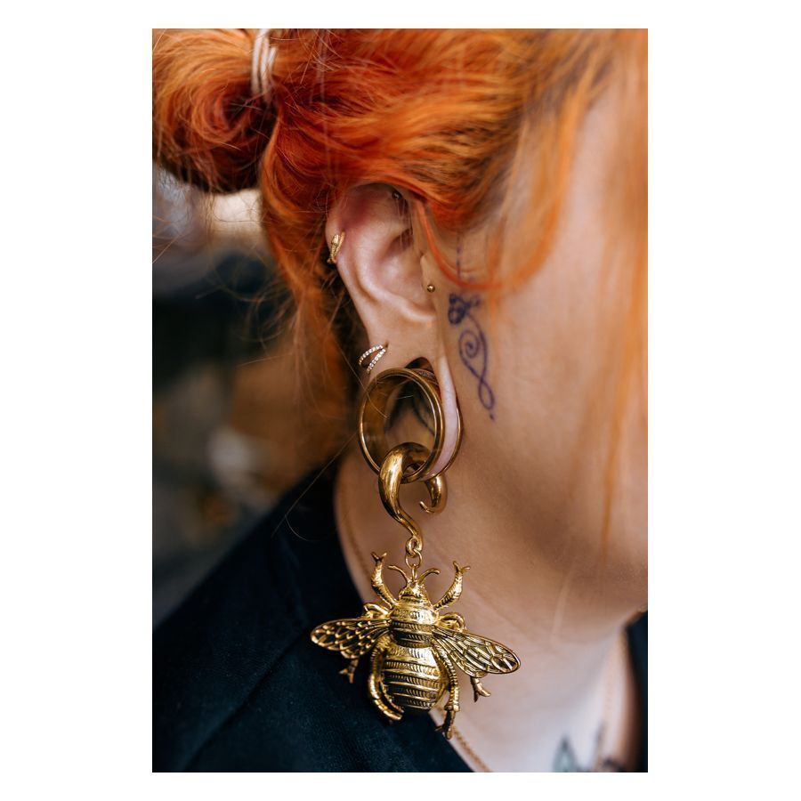 Frau mit rotorangenen Haaren trägt goldenen Ohrring mit großem Bienenanhänger vor verschwommenem Hintergrund