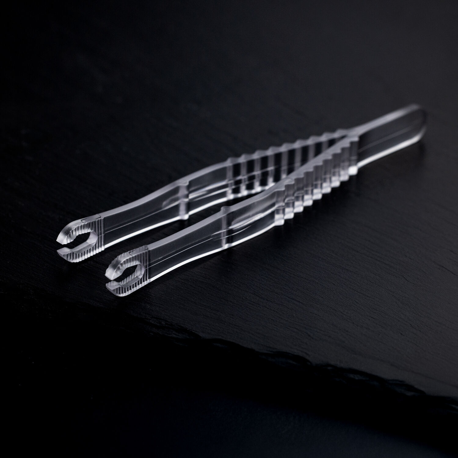 Zwei transparente, sterile Runde Piercingzangen liegen überkreuzt auf einem dunklen Hintergrund.