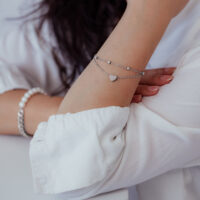 Silbernes zweireihiges Armband mit Herzanhänger am Arm einer Person in weißer Kleidung