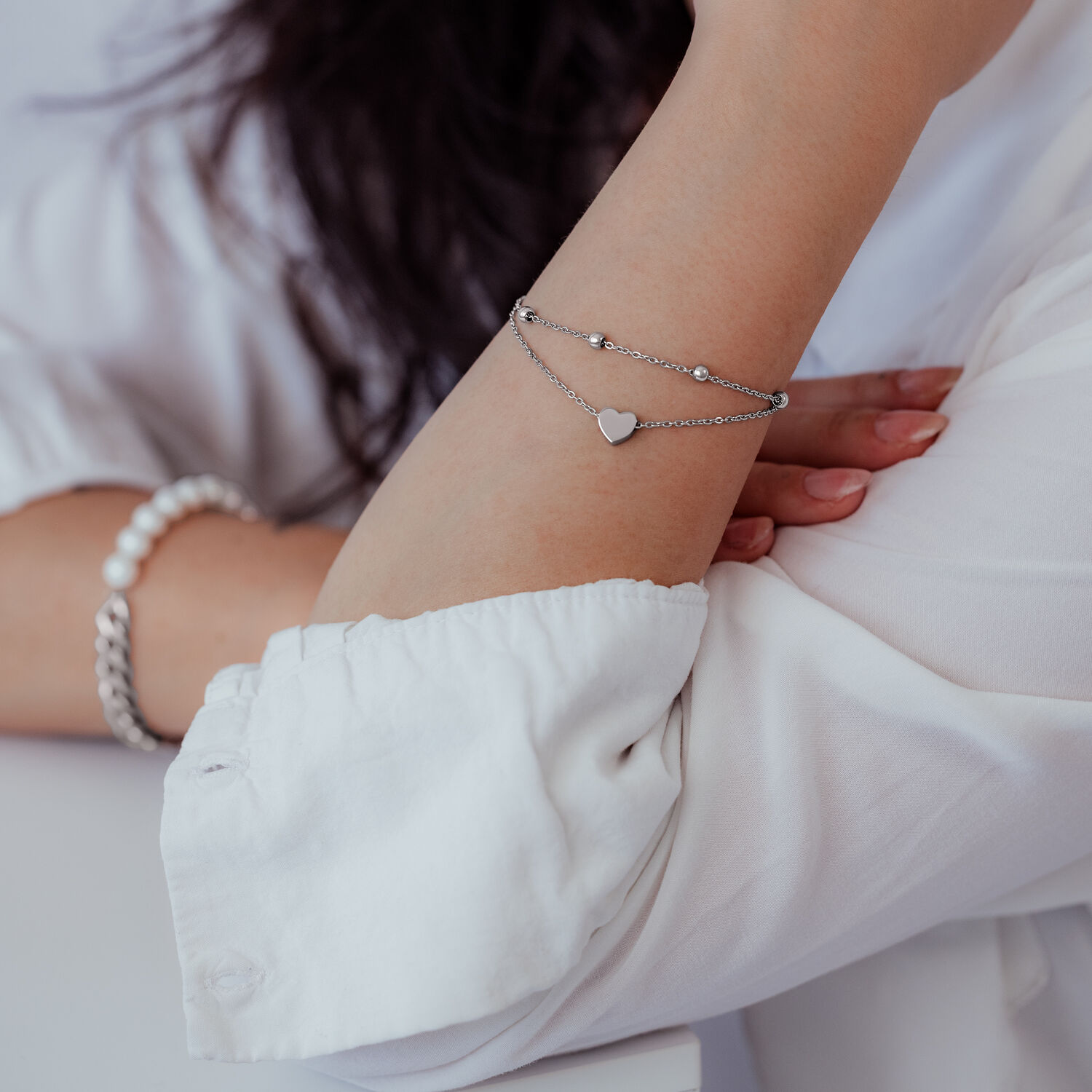 Silbernes zweireihiges Armband mit Herzanhänger am Arm einer Person in weißer Kleidung